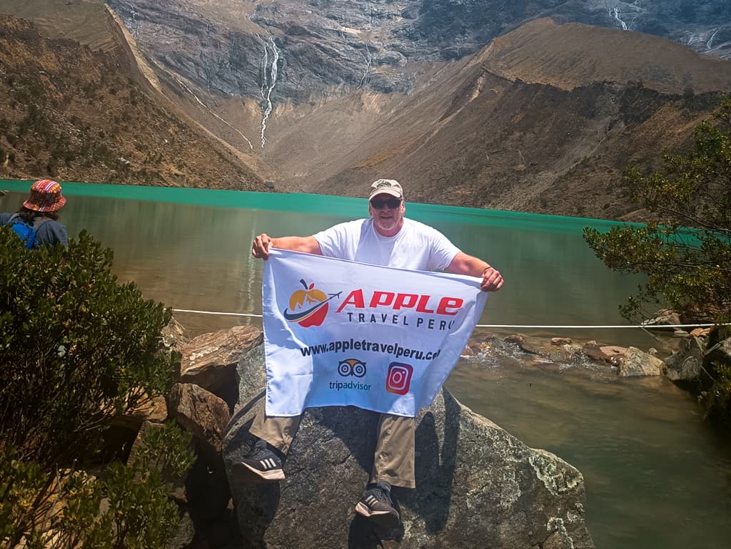 A smiling male tourist with Apple Travel Peru's flag at Humantay Lake
