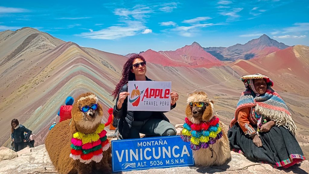 A female tourist with Apple Travel Peru's placard sitting with alpacas and a local at Rainbow Mountain Tour