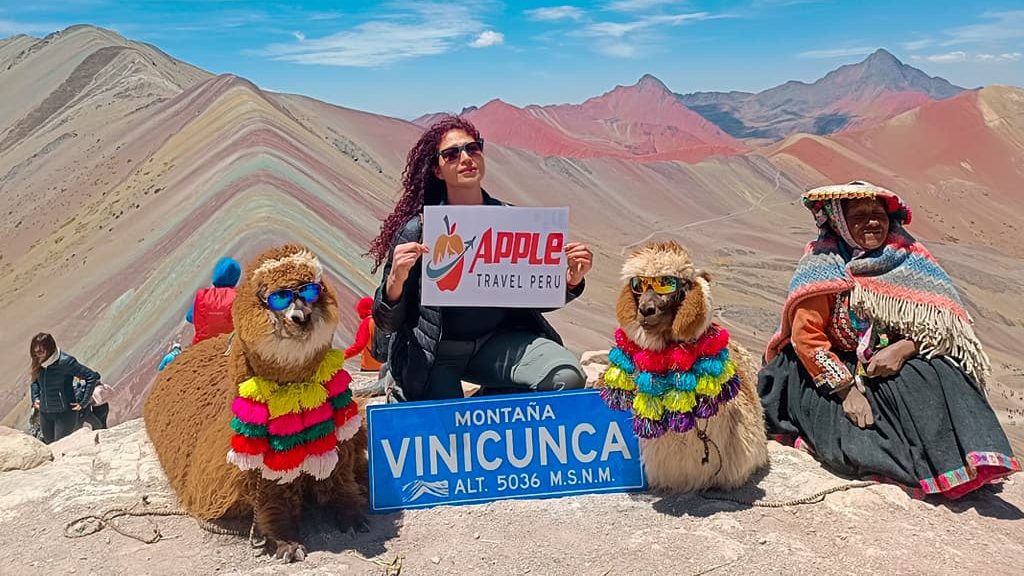 A female tourist with Apple Travel Peru's placard sitting with alpacas and a local at Rainbow Mountain Tour